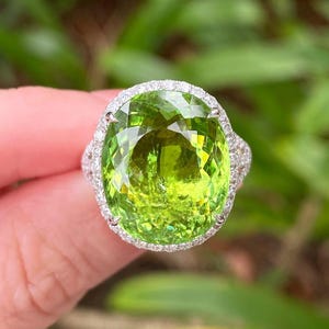 Puede incluir: Un anillo con una gran piedra preciosa verde lima de talla ovalada, rodeada por un halo de pequeñas piedras claras, engastada en una banda plateada. La piedra central del anillo está facetada, reflejando la luz.