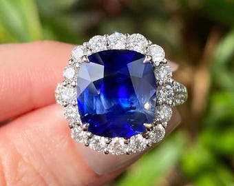 Peut inclure: Gros plan sur une bague ornée d'une grosse pierre précieuse bleue carrée, entourée d'un halo de petites pierres précieuses claires. Le corps de la bague est également orné de petites pierres précieuses claires, créant un effet scintillant. La bague est en métal argenté ou blanc.