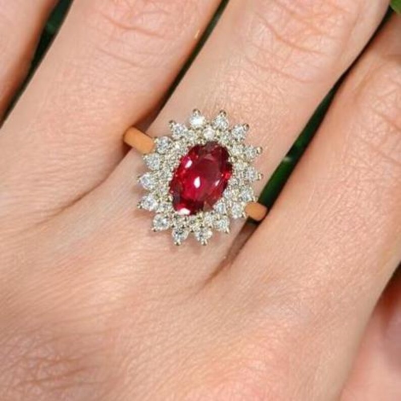 May include: A gold ring featuring a central oval-cut deep red gemstone surrounded by a halo of small, round, clear white stones. The setting is intricate, with the clear stones arranged in a floral-like pattern around the central red stone.