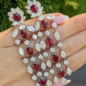 Puede incluir: Un par de pendientes largos colgantes con rub&iacute;es de color rojo intenso en talla ovalada y diamantes transparentes en talla marquesa y redonda, engastados en metal plateado. El dise&ntilde;o presenta m&uacute;ltiples hileras de gemas en cascada.