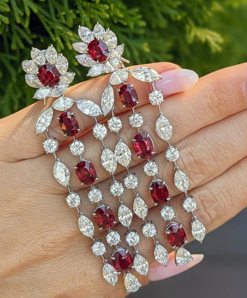 Puede incluir: Un par de pendientes largos colgantes con rub&iacute;es de color rojo intenso en talla ovalada y diamantes transparentes en talla marquesa y redonda, engastados en metal plateado. El dise&ntilde;o presenta m&uacute;ltiples hileras de gemas en cascada.