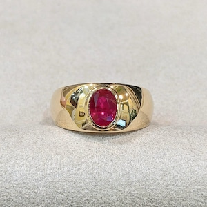 May include: A polished yellow gold ring featuring a central oval-cut vibrant red ruby gemstone. The ruby is securely set within a smooth, wide, tapered gold band that reflects light across its curved, high-shine metallic surface.