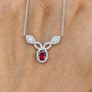 May include: A delicate silver-toned necklace featuring a central oval-cut deep red gemstone surrounded by a halo of small clear stones. The pendant design extends into two symmetrical branches, each accented with additional clear stones on a chain.