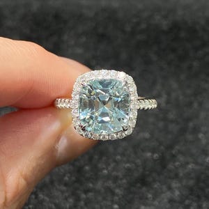 Puede incluir: Un anillo de plata con una gran piedra preciosa de talla cojín azul claro, rodeada por un halo de piedras claras más pequeñas. La banda del anillo también está adornada con pequeñas piedras claras. El anillo es sostenido por una mano sobre un fondo oscuro.