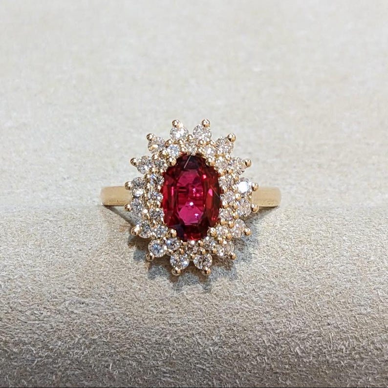 May include: A gold ring featuring a central oval-cut deep red gemstone surrounded by a halo of small, round, clear white diamonds. The setting is intricate, with the diamonds arranged in a floral-like pattern around the vibrant red stone.