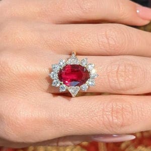 K&ouml;nnte beinhalten: Ein Ring mit einem gro&szlig;en, oval geschliffenen, tiefroten Edelstein in der Mitte, umgeben von einem Kranz aus kleineren, klaren, rund geschliffenen Steinen in einer Metallschiene. Das Design bildet eine bl&uuml;tenartige Anordnung um den roten Stein.