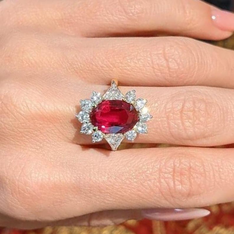 K&ouml;nnte beinhalten: Ein Ring mit einem gro&szlig;en, oval geschliffenen, tiefroten Edelstein in der Mitte, umgeben von einem Kranz aus kleineren, klaren, rund geschliffenen Steinen in einer Metallschiene. Das Design bildet eine bl&uuml;tenartige Anordnung um den roten Stein.