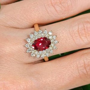 May include: A gold ring featuring a central oval-cut deep red gemstone surrounded by a halo of numerous small, clear, round-cut white stones arranged in a starburst pattern, set on a thin, polished gold band.