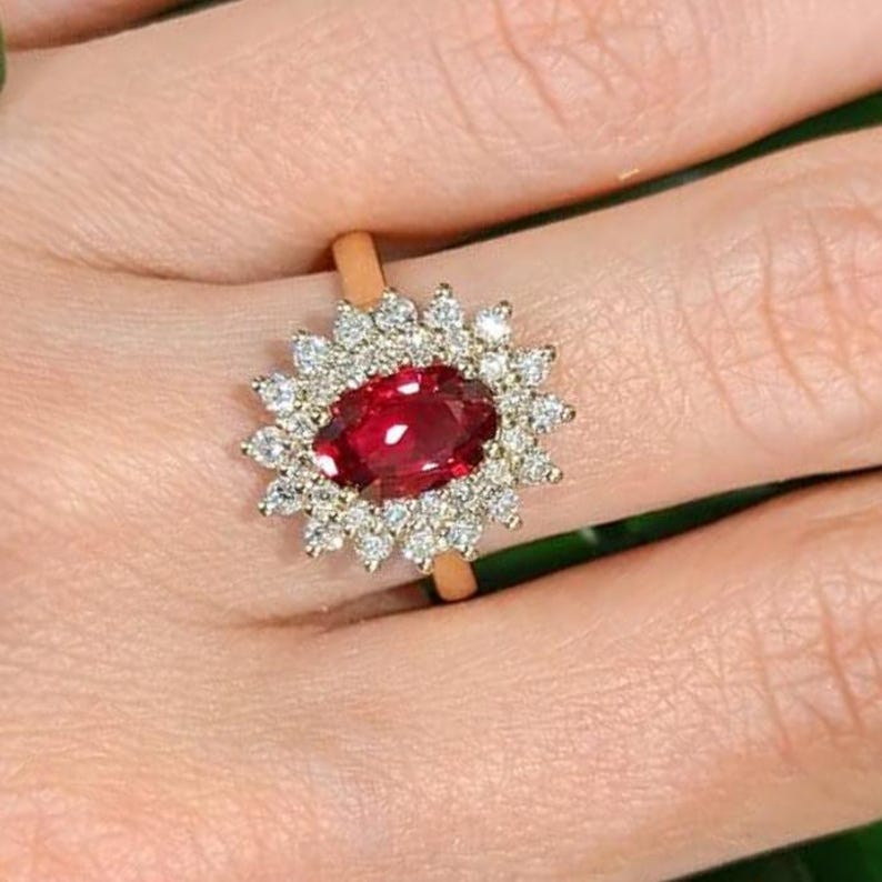 May include: A gold ring featuring a central oval-cut deep red gemstone surrounded by a halo of numerous small, clear, round-cut white stones arranged in a starburst pattern, set on a thin, polished gold band.