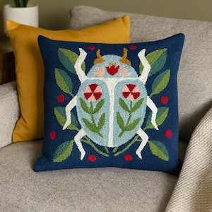 Op de afbeelding: Een marineblauw sierkussen met een gedetailleerd geborduurd ontwerp van een gestileerde kever. De kever heeft een lichtblauw lichaam met rode en roze bloemaccenten, witte poten en is omgeven door groene bladeren en rode stippen.