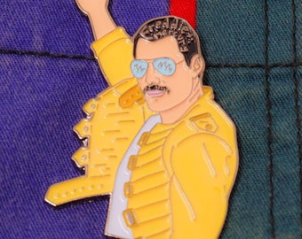 May include: A yellow enamel pin featuring a cartoon illustration of Freddie Mercury wearing a yellow jacket and sunglasses, with his arm raised in the air. The pin is on a blue and green striped fabric background.