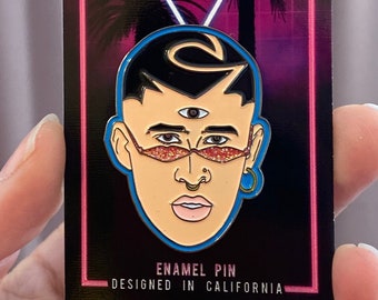 May include: A pink and blue enamel pin featuring a cartoon portrait of a man with a third eye and pink glitter sunglasses. The pin is on a card with the text "Old School Pins" and "Enamel Pin Designed in California".