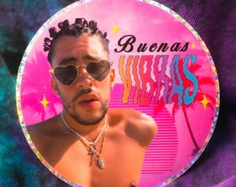 May include: A round sticker with a pink background and a holographic finish. The sticker features a photo of a man with dark hair and a beard wearing heart-shaped sunglasses. The text "Buenas Vibras" is written in a colorful, stylized font.