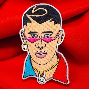 May include: Embroidered patch featuring a cartoon portrait of a person with black hair, pink sunglasses, a gold chain, and a blue and red shirt. The person is wearing a nose ring and a small bunny charm.