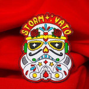 May include: Embroidered patch featuring a stylized Stormtrooper helmet design, incorporating elements of a sugar skull. The patch is white with colorful accents, including red roses, yellow stars, and the text "STORM VATO" in yellow.