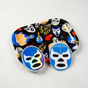 May include: A black tray with a colorful lucha libre mask design, featuring the text "Lucha Now". Two lucha libre mask patches, one blue and white, the other iridescent blue, are placed on the tray.