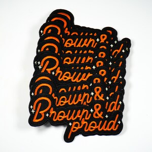 May include: A stack of black iron-on patches with white stitching. The patches feature the text "Brown & Proud" in orange embroidery.