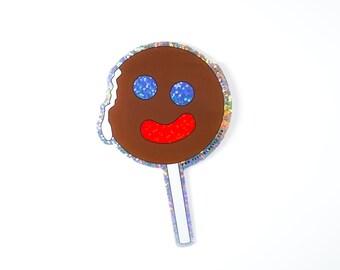 May include: A brown gingerbread man lollipop with a white icing swirl and a smiling face with blue eyes and a red mouth. The lollipop is on a white stick.