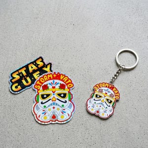 May include: A set of stickers and a keychain featuring a stylized Stormtrooper helmet design. The helmet is decorated with colorful floral and Day of the Dead-inspired elements, with the text "STAS GUEY" and "STORM + VATO".