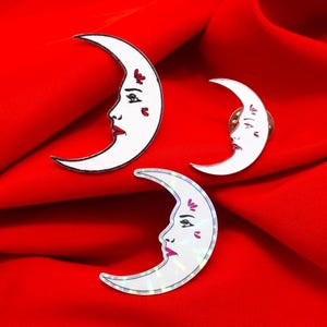 May include: Three crescent moon pins and patches with a face design. The moons are white with red lips and accents. One is a patch, one is a pin, and one has a holographic finish. The background is a red fabric.