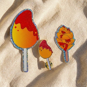 May include: Three colorful enamel pins shaped like mango treats. The pins feature a vibrant gradient of yellow and orange, topped with red sauce. The pins have a silver glittery border and are set against a neutral background.