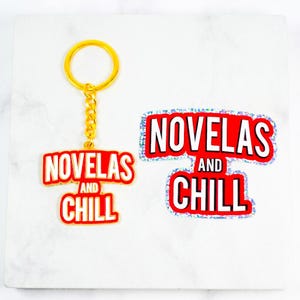 May include: A gold-colored keychain and sticker set with the words "NOVELAS AND CHILL" in red and white. The keychain has a gold ring and chain. The sticker has a glittery border.