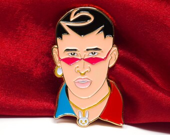 May include: Gold-colored enamel pin featuring a cartoon portrait of a man with black hair, pink sunglasses, a gold chain with a white rabbit pendant, and a blue and red shirt.