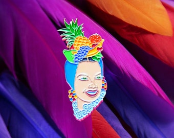May include: A colorful enamel pin featuring a woman with a blue headscarf and a fruit headdress. The pin is set against a background of colorful feathers.