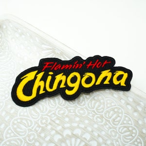 May include: Embroidered patch with the words "Flamin' Hot Chingona" in red and yellow thread on a black background. The patch has a curved shape and is likely designed to be ironed or sewn onto clothing or accessories.