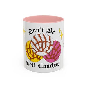 May include: A white ceramic mug with a pink interior rim featuring a graphic of three concha sweet breads in yellow, white, and pink. The text Don't Be Self-Conchas is printed in black gothic lettering above and below the breads with yellow stars.