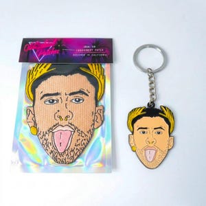 May include: An iron-on embroidery patch and a keychain featuring a stylized face with a gold laurel crown, gold earring, and tongue sticking out. The patch is in a holographic package with text that reads "Outlandish Patches, Iron-On Embroidery Patch, Designed in California."