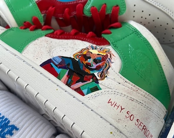 May include: White sneakers with green and red accents. The sneakers have a Joker-themed design with a colorful portrait of the Joker and the text "WHY SO SERIOUS?" on the side.