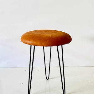 Könnte beinhalten: Ein runder, brauner Lederhocker mit schwarzen Haarnadelbeinen. Der Sitz des Hockers hat eine warme, karamellfarbene Farbe, und die Beine sind dünn und abgewinkelt, wodurch eine moderne Ästhetik der Mitte des Jahrhunderts entsteht. Der Hocker ist ein einfaches, aber stilvolles Möbelstück.