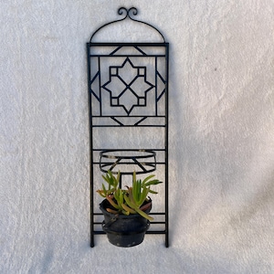 May include: Black metal wall planter with a geometric design and a small black pot holding a succulent plant. The planter has a decorative top and a circular holder for the pot. The plant has green and red leaves.