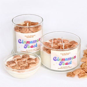 May include: Three glass jars filled with a white soy candle and cinnamon toast cereal pieces. The jars have labels that read "Cinnamon Toast" and "Good By Summer Candle Co.". There are also loose cinnamon toast cereal pieces scattered on a white surface.