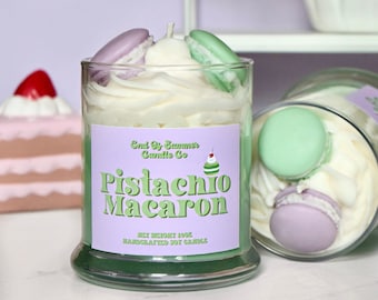 May include: A clear glass jar candle with a green and purple label that reads "Pistachio Macaron". The candle is topped with white wax and decorative macarons. A second candle is visible, along with a decorative shelf and cake.