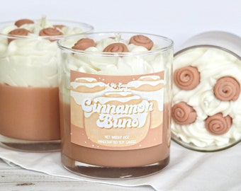 May include: Three glass jars of cinnamon bun scented soy candles. The candles are topped with a white frosting and brown cinnamon bun swirls. The label on the middle jar reads "End of Summer Candle Co. Cinnamon Buns. Net Weight 10oz. Handcrafted Soy Candle."