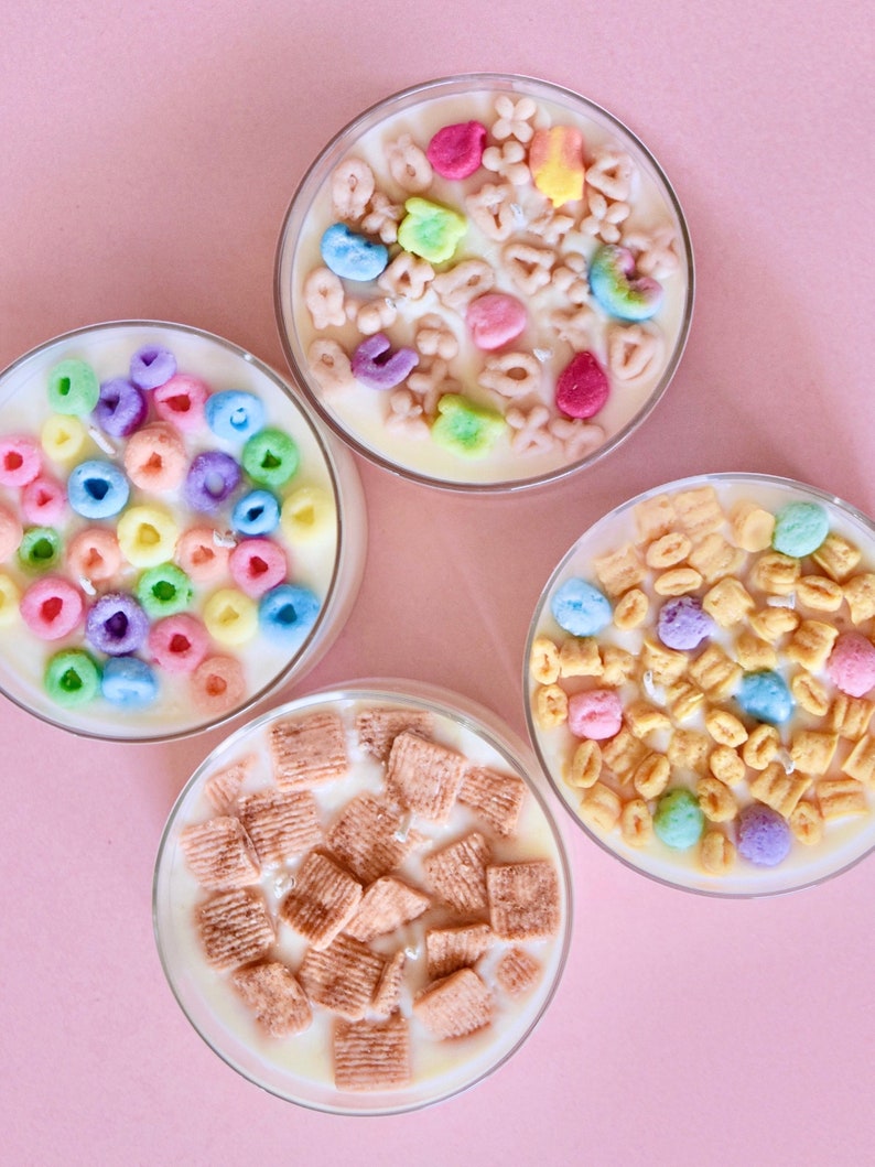 May include: Four glass candle jars with different cereal-themed designs. The candles are white with various colorful cereal pieces embedded in the wax. One jar features colorful cereal rings, another has colorful letter-shaped cereal, a third has small, colorful cereal pieces, and the last jar has brown, square-shaped cereal pieces.