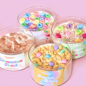 May include: Four glass jars filled with colorful cereal-themed soy candles. The candles are labeled "Cinnamon Toast", "Cereal Milk", "Magic Charms", and "Crunch Berry".