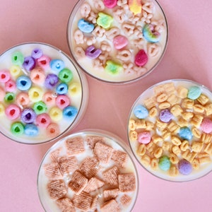 May include: Four glass jars filled with white wax and topped with different types of cereal. The jars are on a pink background.