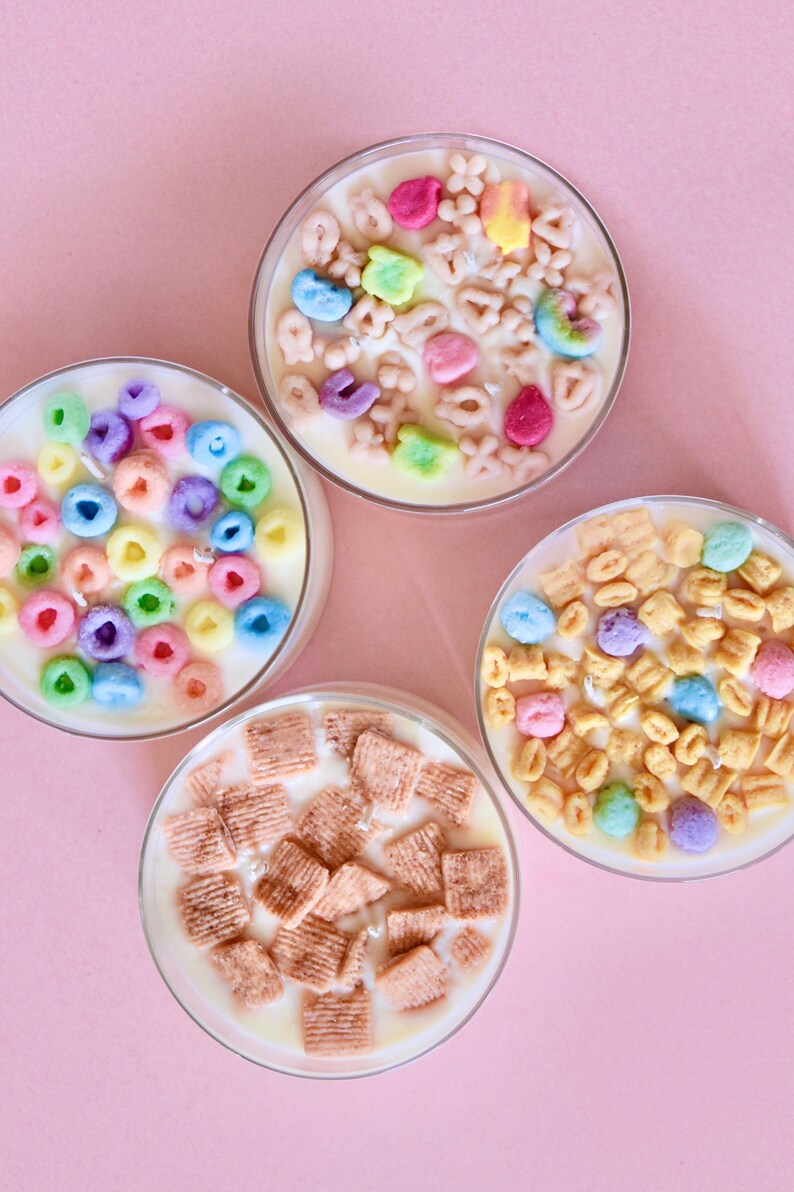 May include: Four glass jars filled with white wax and topped with different types of cereal. The jars are on a pink background.