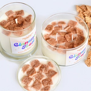 May include: Three glass jars filled with a white candle and cinnamon toast cereal pieces. The jars have labels that say "Cinnamon Toast" in colorful letters.