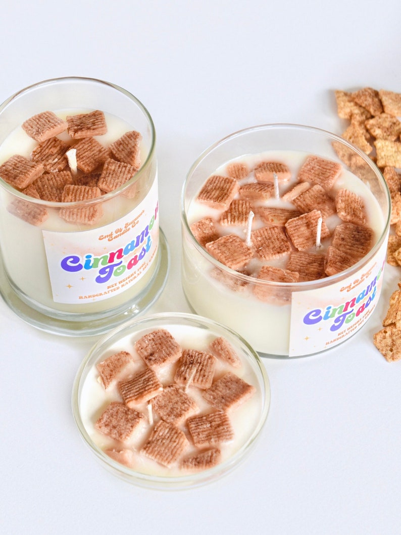 May include: Three glass jars filled with a white candle and cinnamon toast cereal pieces. The jars have labels that say "Cinnamon Toast" in colorful letters.