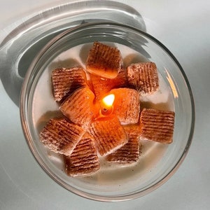 May include: A clear glass jar filled with a white substance and topped with a cluster of brown, cinnamon-scented wax squares. A lit candle sits in the center of the wax squares.