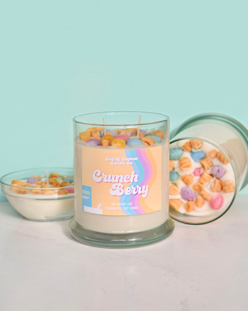 May include: A glass jar candle with a white label that says "Crunch Berry" and "End of Summer Candle Co.". The candle is topped with colorful cereal pieces.
