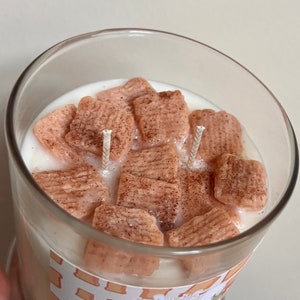 May include: A glass jar candle with a white wax base and a cinnamon-scented, pink cereal-shaped wax topping. The candle has two wicks.