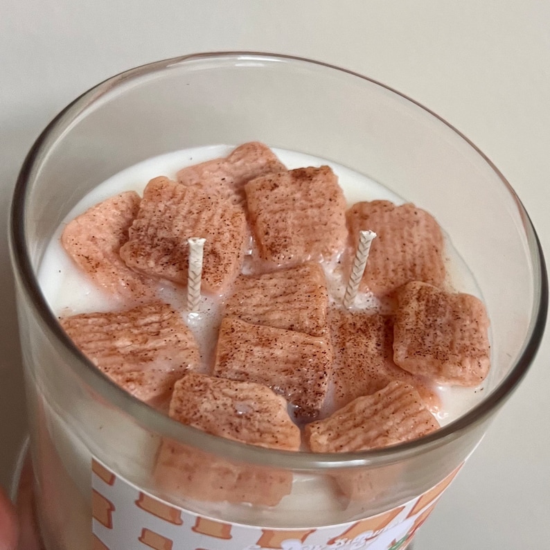May include: A glass jar candle with a white wax base and a cinnamon-scented, pink cereal-shaped wax topping. The candle has two wicks.