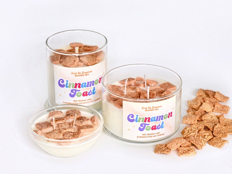 May include: Three glass jars filled with a white soy candle and cinnamon toast cereal pieces. The jars have labels that read "Cinnamon Toast" and "Good By Summer Candle Co.". There are also loose cinnamon toast cereal pieces scattered on a white surface.