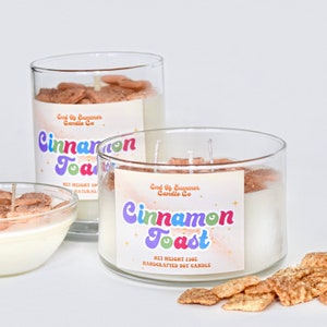 May include: Three glass jars of cinnamon toast candle with a label that reads "End of Summer Candle Co. Cinnamon Toast Net Weight 13oz Handcrafted Soy Candle". The candle jars are filled with white wax and topped with cinnamon toast cereal. There are also cinnamon toast cereal pieces scattered on the surface.