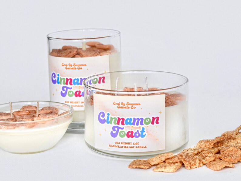May include: Three glass jars of cinnamon toast candle with a label that reads "End of Summer Candle Co. Cinnamon Toast Net Weight 13oz Handcrafted Soy Candle". The candle jars are filled with white wax and topped with cinnamon toast cereal. There are also cinnamon toast cereal pieces scattered on the surface.